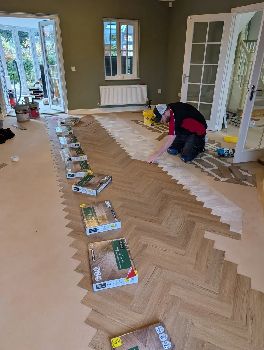 Marc kneeling on the floor laying warm oak herringbone planks, with boxes of flooring stacked in a diagonal line across the room.