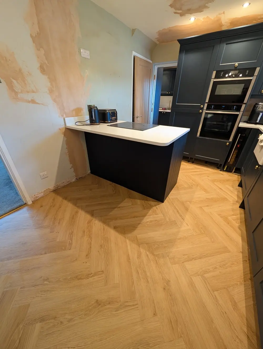 Natural oak herringbone LVT running across a kitchen floor past a dark navy island unit.