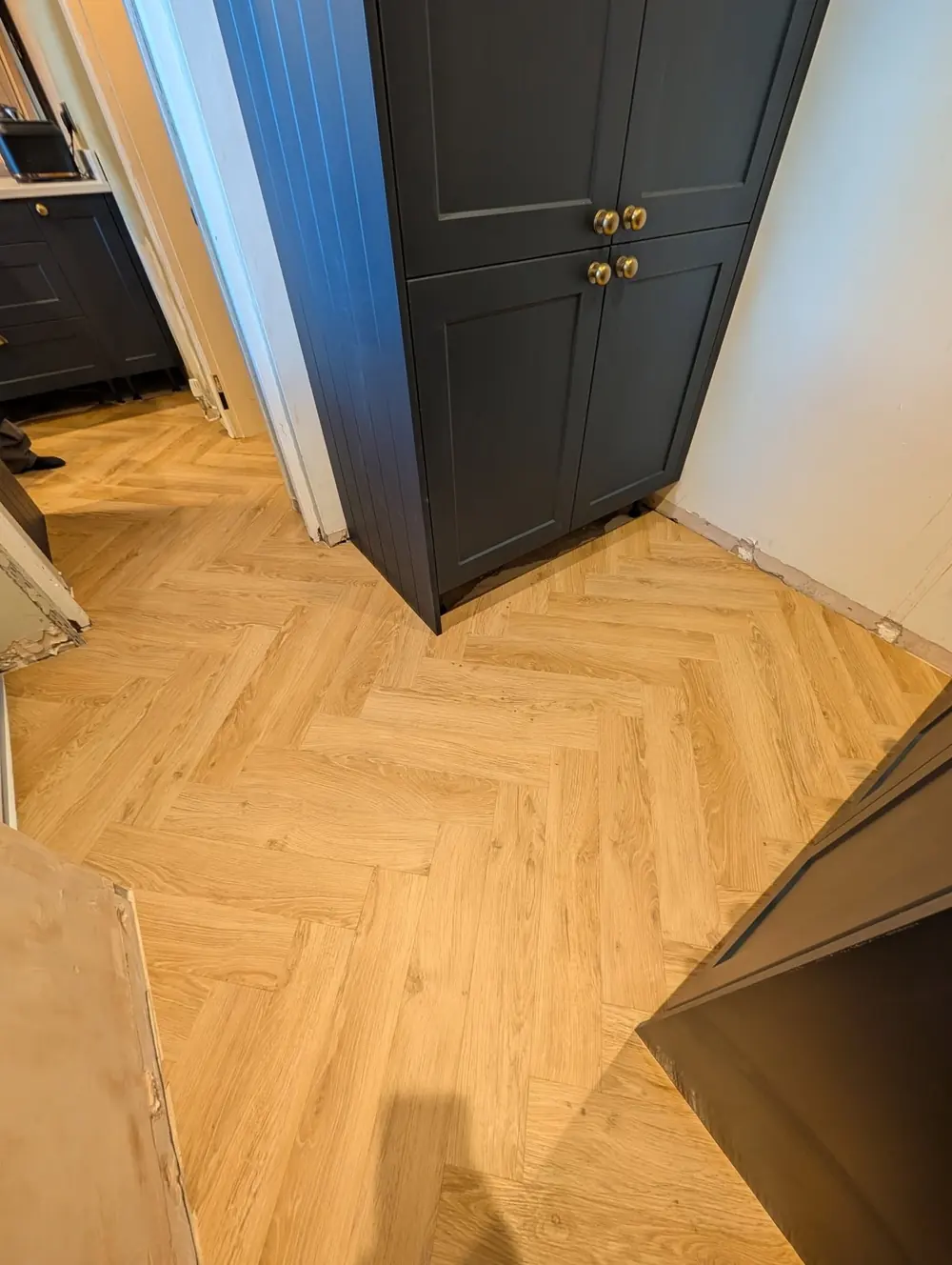 Natural oak herringbone meeting the corner of a navy larder unit with brass cup handles.