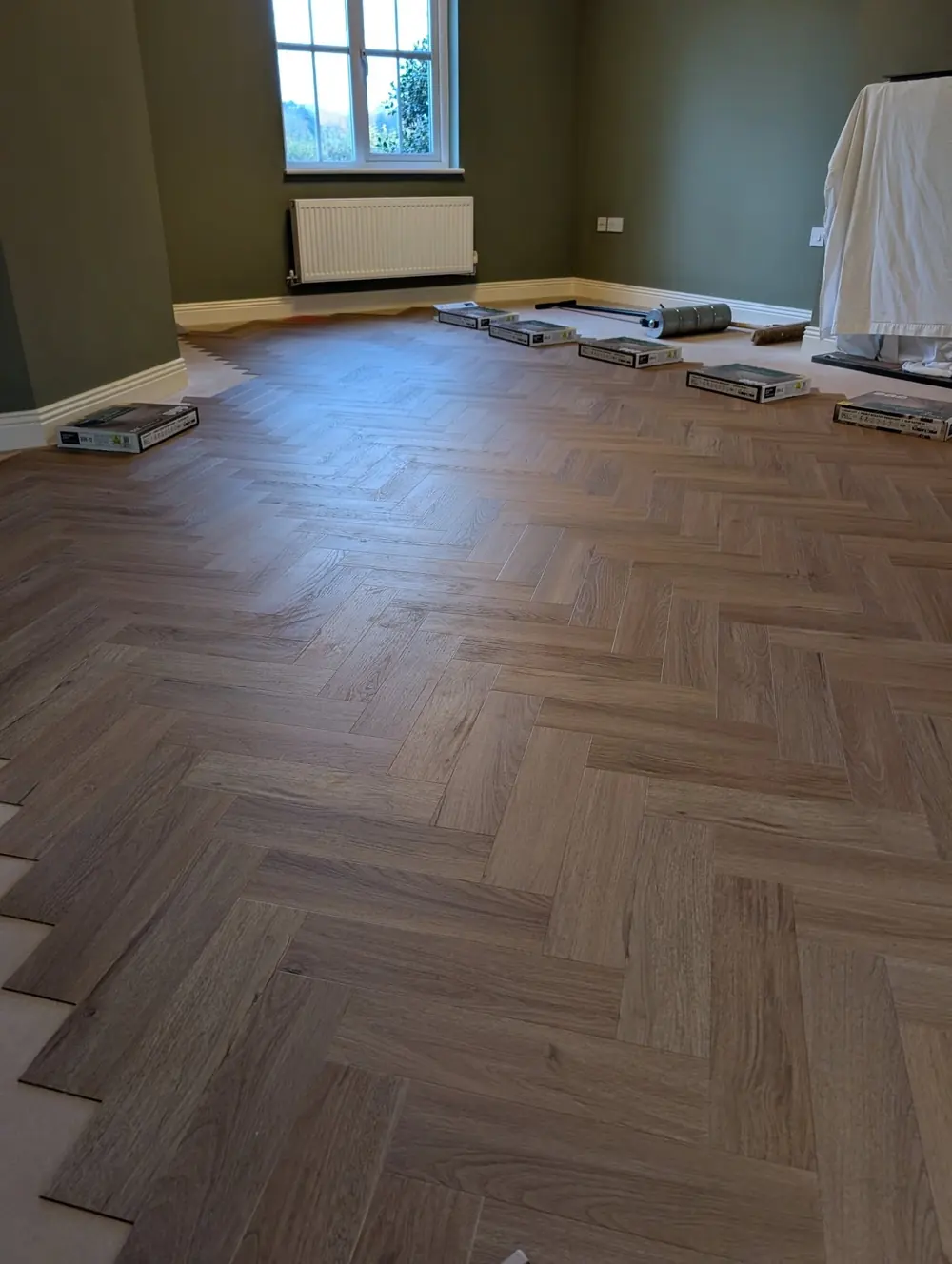 Warm oak herringbone LVT filling a large olive green room with a window and dust sheets to one side.