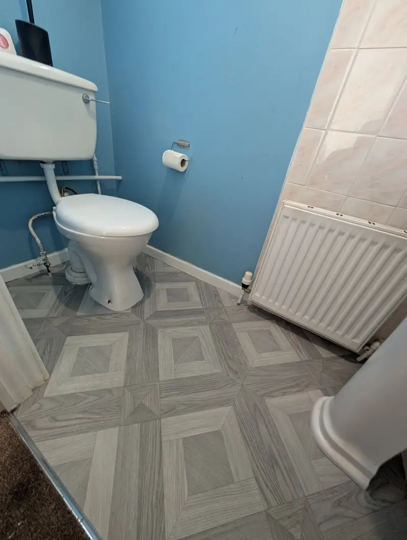 Patterned vinyl in a blue walled cloakroom, cut around a toilet pan and under a radiator.